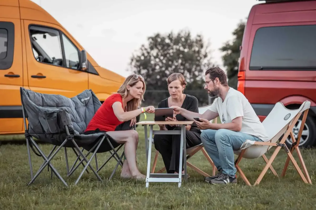 Jenny und Lukas von der Camping App sitzen auf Camping-Stühlen vor zwei Vans und erklären einer Nutzerin die App