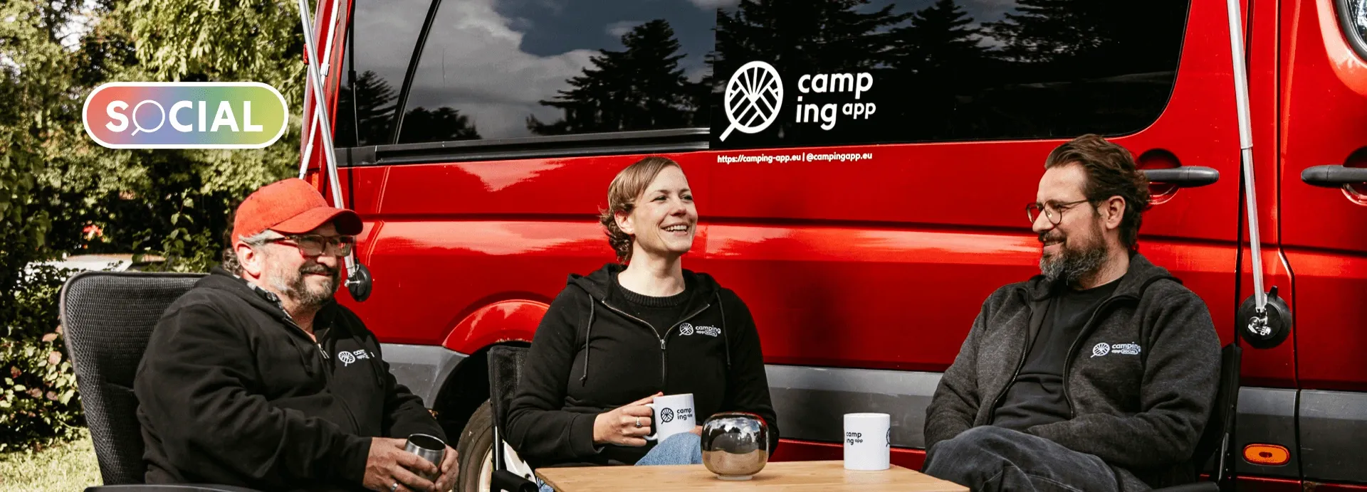 Jenny Lukas und Dirk von der Camping App Social sitzen lachend vor einem roten Camper