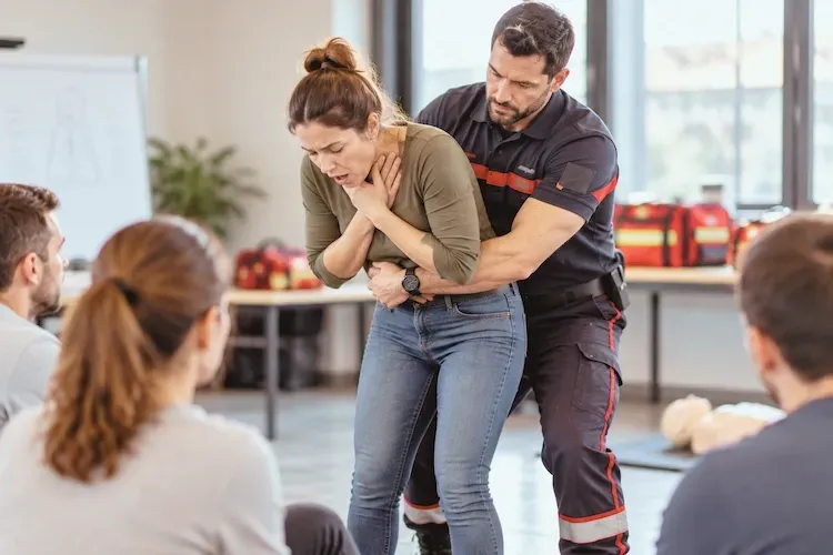 Le formateur réalise une compression abdominale sur une participante qui porte les mains à sa gorge, devant le groupe assis en demi-cercle