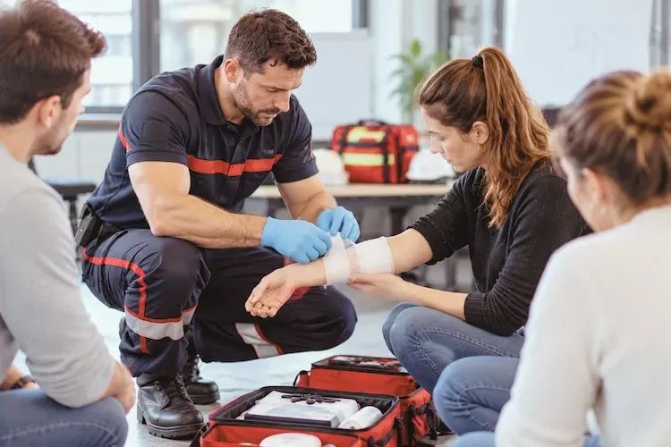 Le formateur, ganté et à genoux, pose un bandage sur l'avant-bras d'une participante assise, sacoche de premiers secours ouverte devant lui