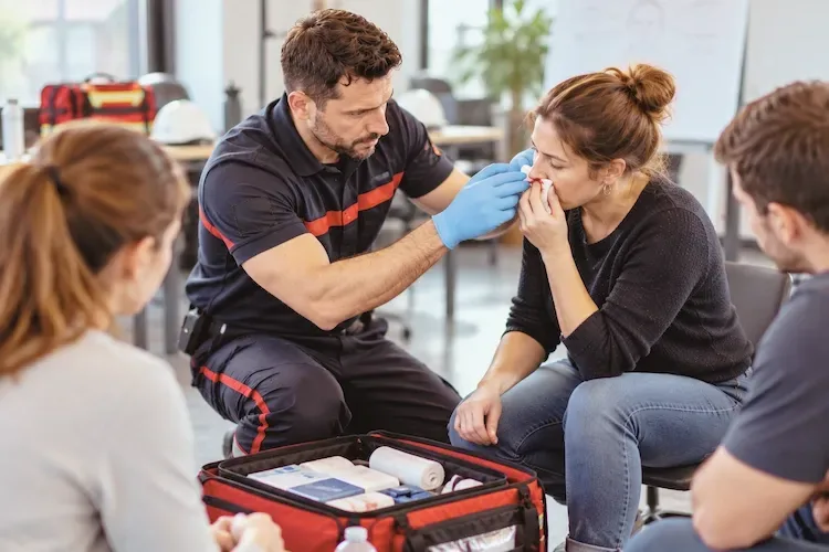Le formateur, ganté, aide une participante à comprimer un saignement de nez avec une compresse, sacoche de premiers secours ouverte au sol