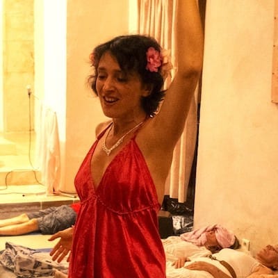 Une femme brune avec une robe rose satinée et une fleur dans les cheveux, chante avec un bras levé pour des personnes allongées sur des matelas, dans une vaste salle en pierre.