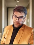 Headshot of a man with dark hair, beard, and eyeglasses, wearing a black turtleneck and golden blazer, looking confidently at the camera, conveying professionalism and approachability.jpeg