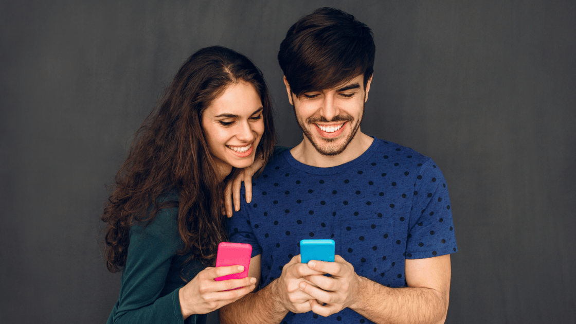 Young couple smiling while looking at their smartphones together against a plain background..png