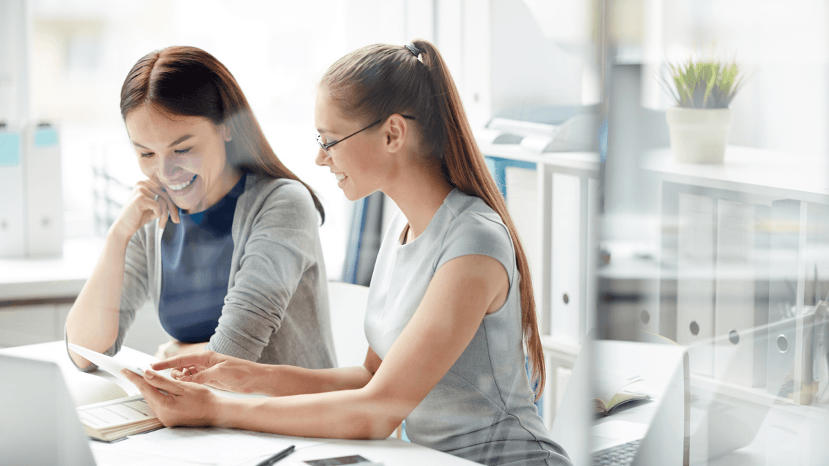 Two women smiling and collaborating with a tablet in a modern office. .png